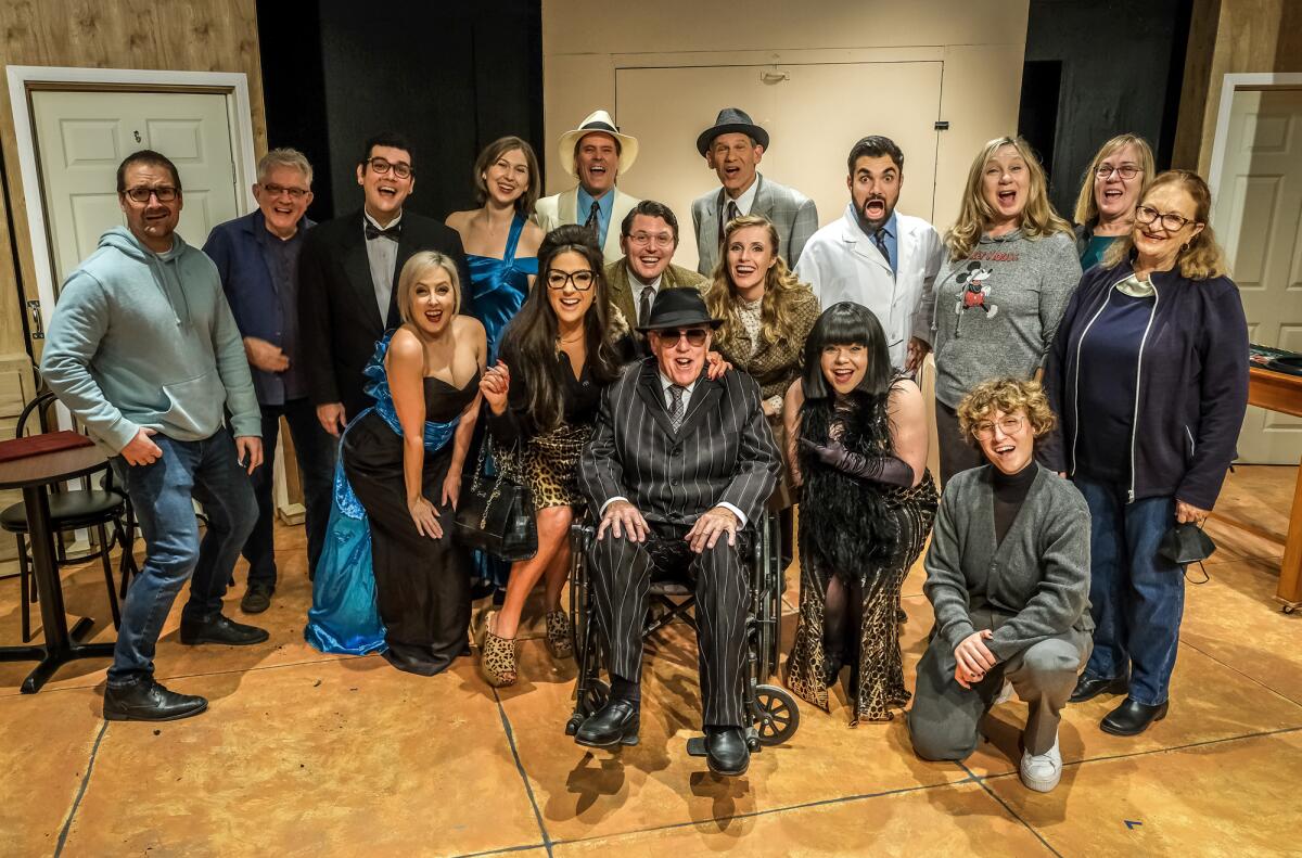 A man wearing a striped suit and hat and sitting in a wheelchair is surrounded by actors and crew members.