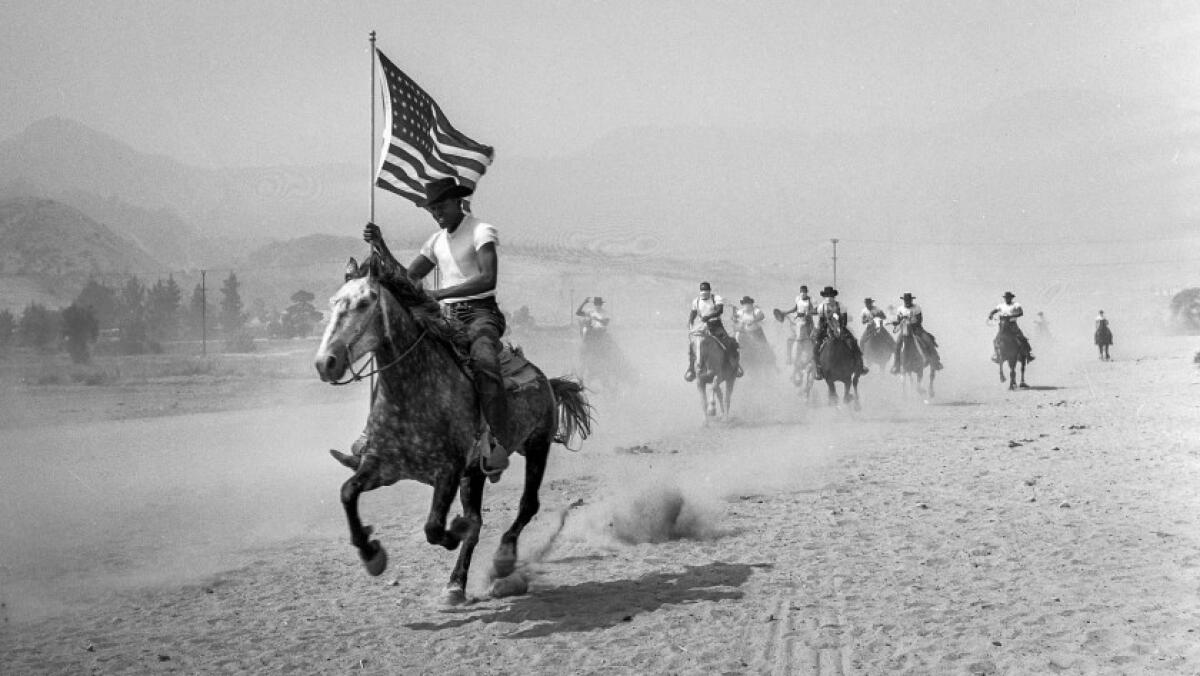 A group of men on horseback riding across a dusty plain, the one in the foreground holding an American flag.