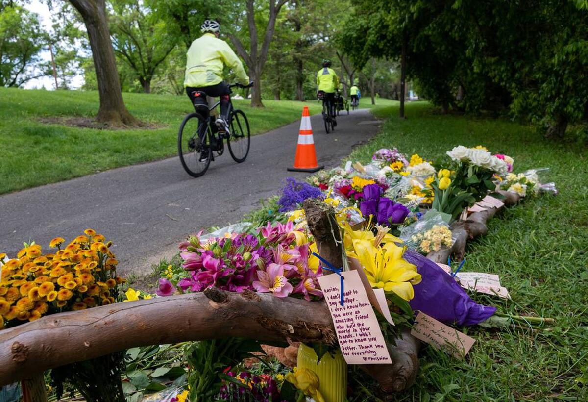 A cyclist rides on a paved path through a grassy park, passing a mound of bouquets and cards on the grass.