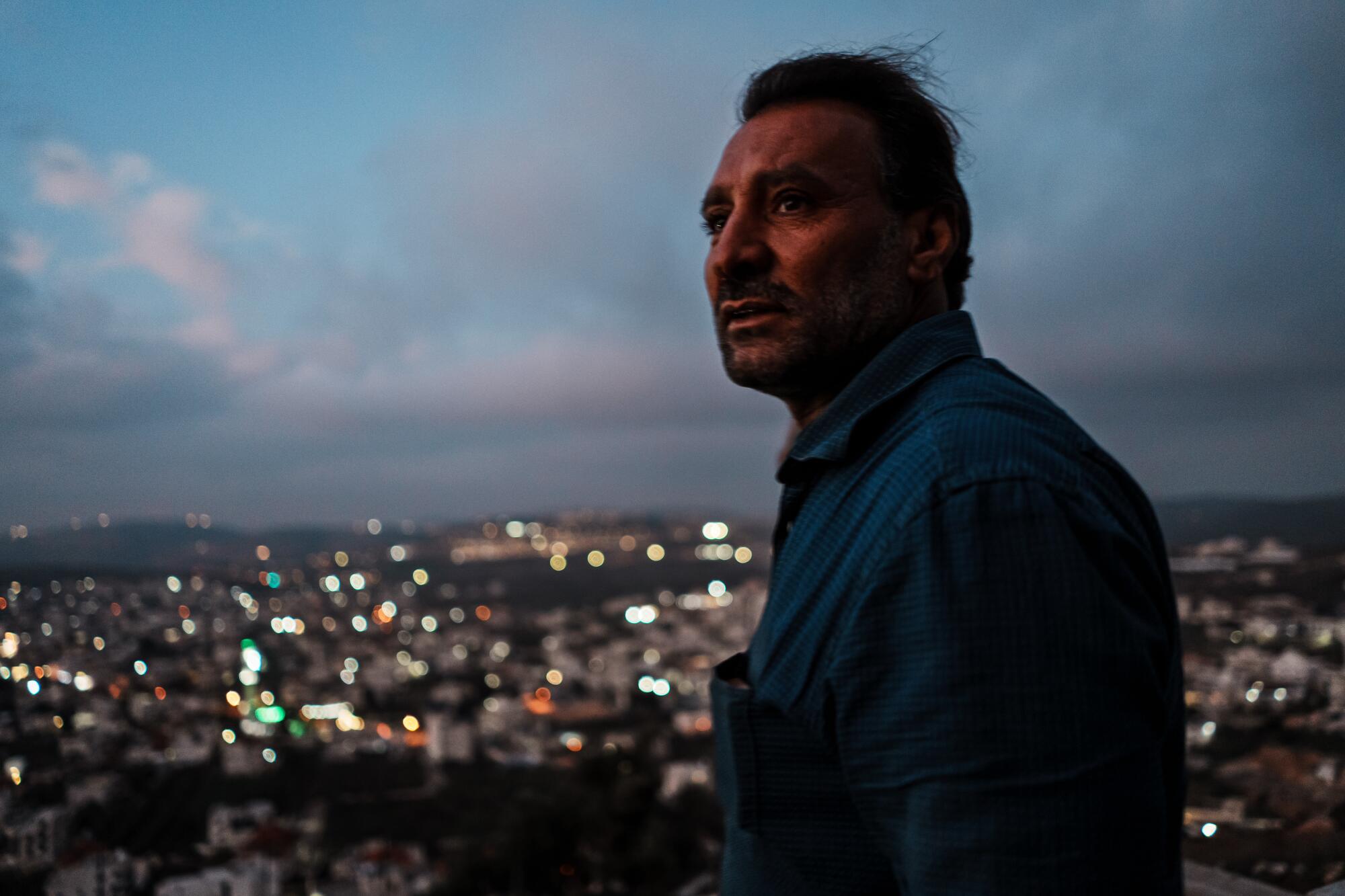 A man pictured from the chest up before nightfall, city lights visible below him in the background