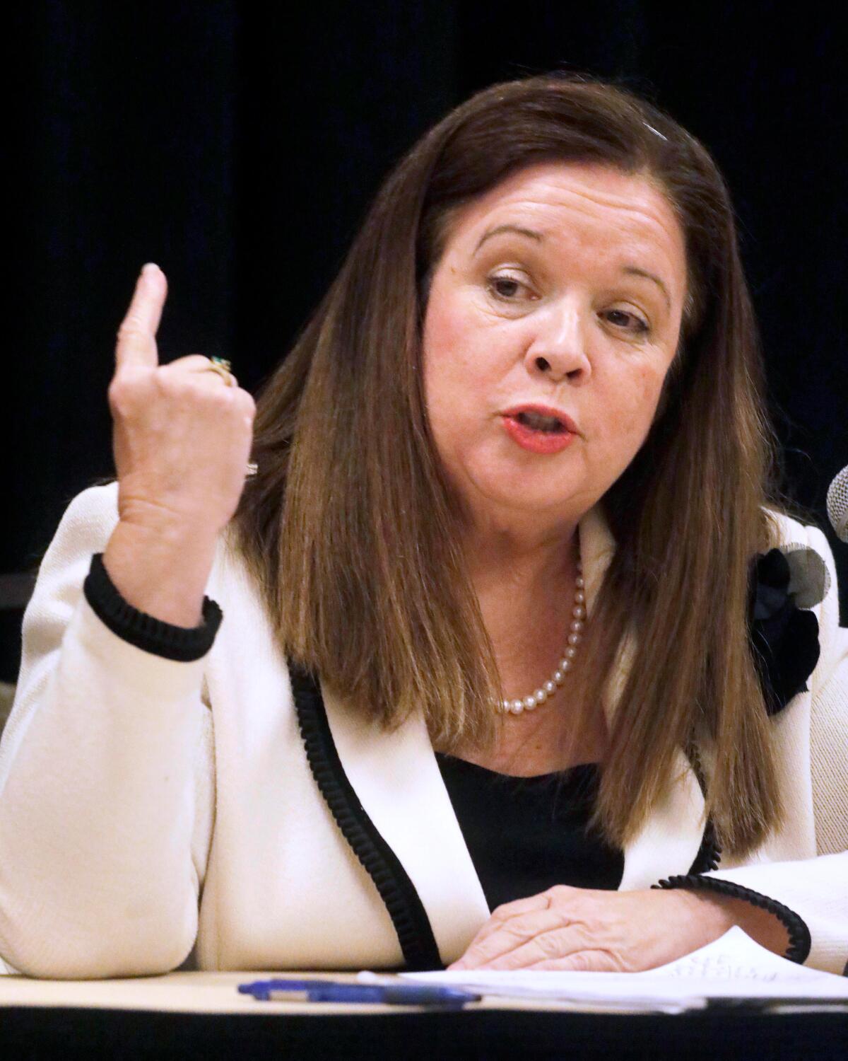 A vertical frame of a woman with medium-length brown hair in a white suit seated, gesturing with right hand while speaking