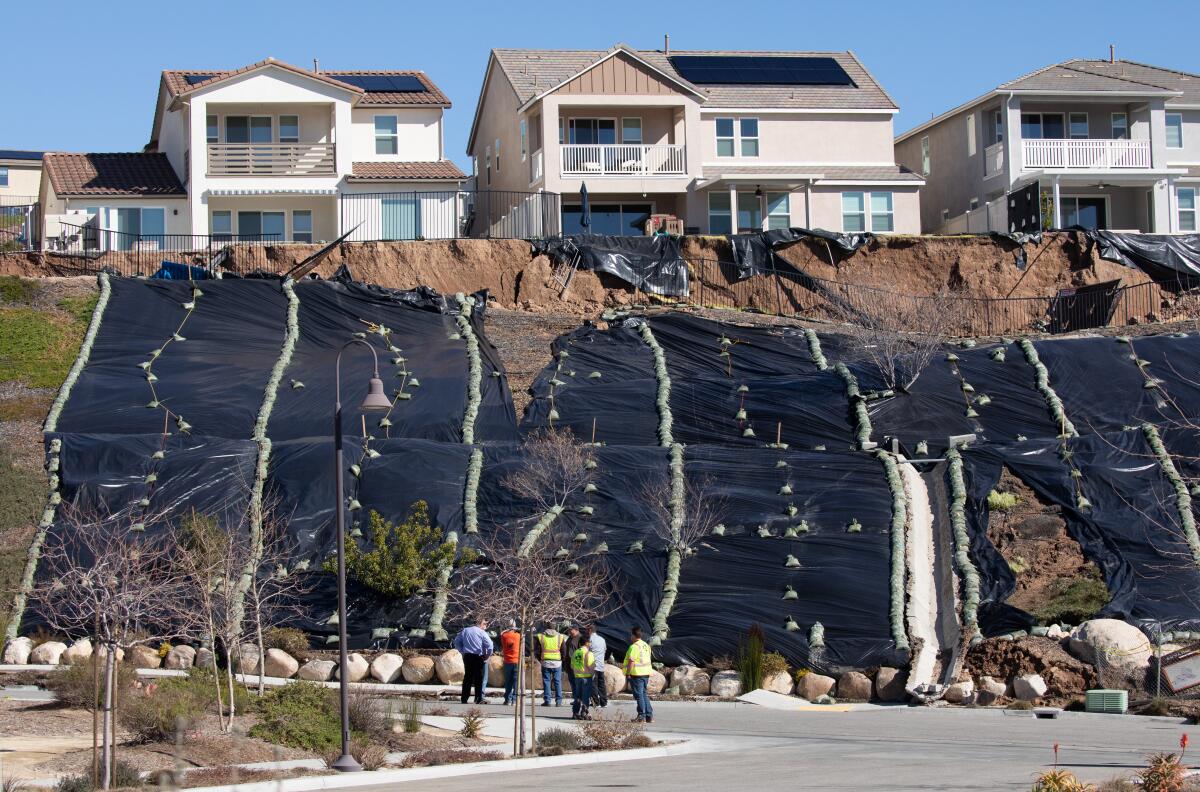 Six people stand at the bottom of a hillside covered with black plastic weighted down with rocks. Three houses are on top.