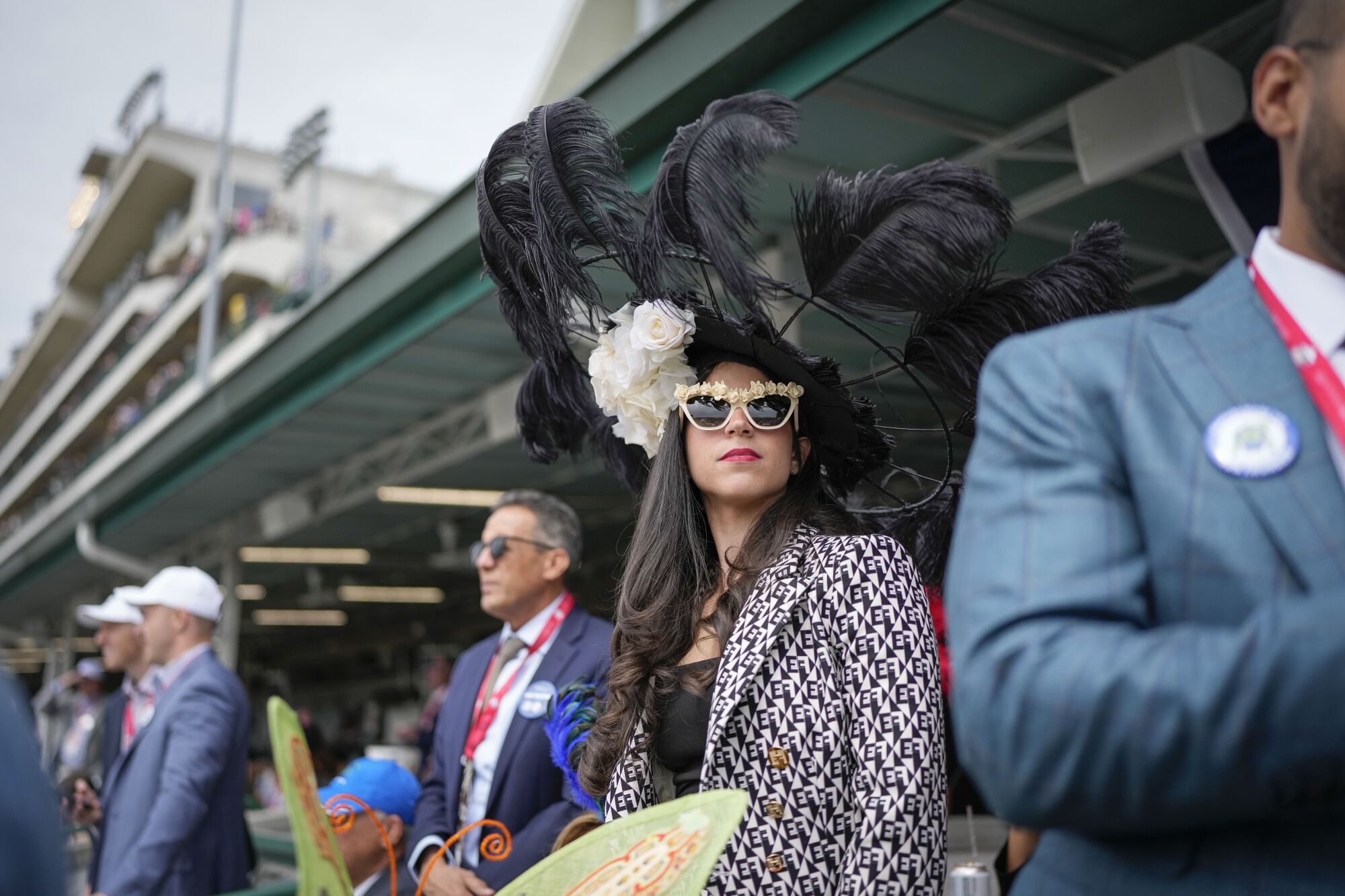 A woman with an elaborate wide-brimmed hat featuring tall black feathers watches a race at Churchill Downs.