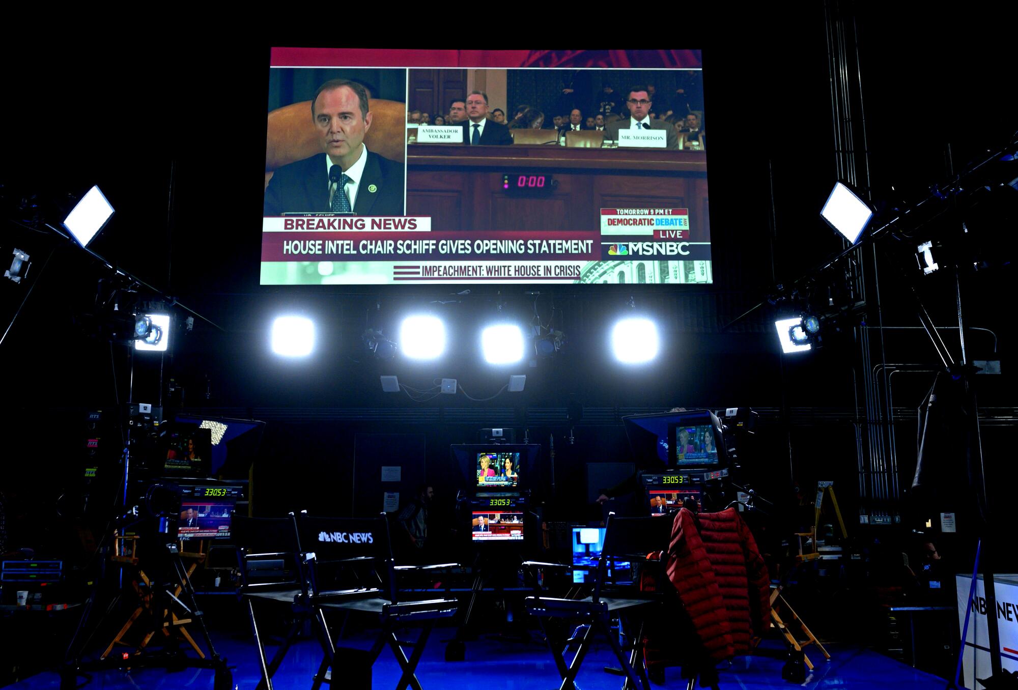 Above stage lights in a dark studio, a large TV reading "breaking news" shows Schiff speaking on a split screen at a hearing