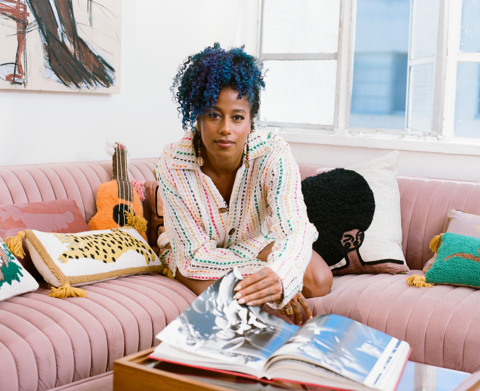 aja monet sits on a pink couch flipping through a large book.