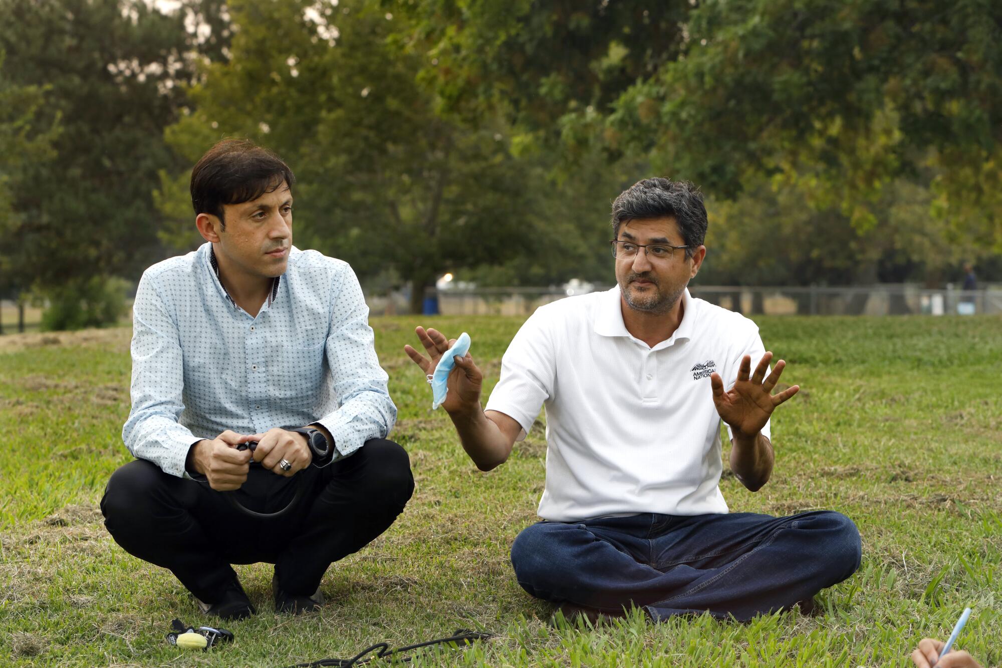 Two men sit in a park, one gesturing while he talks