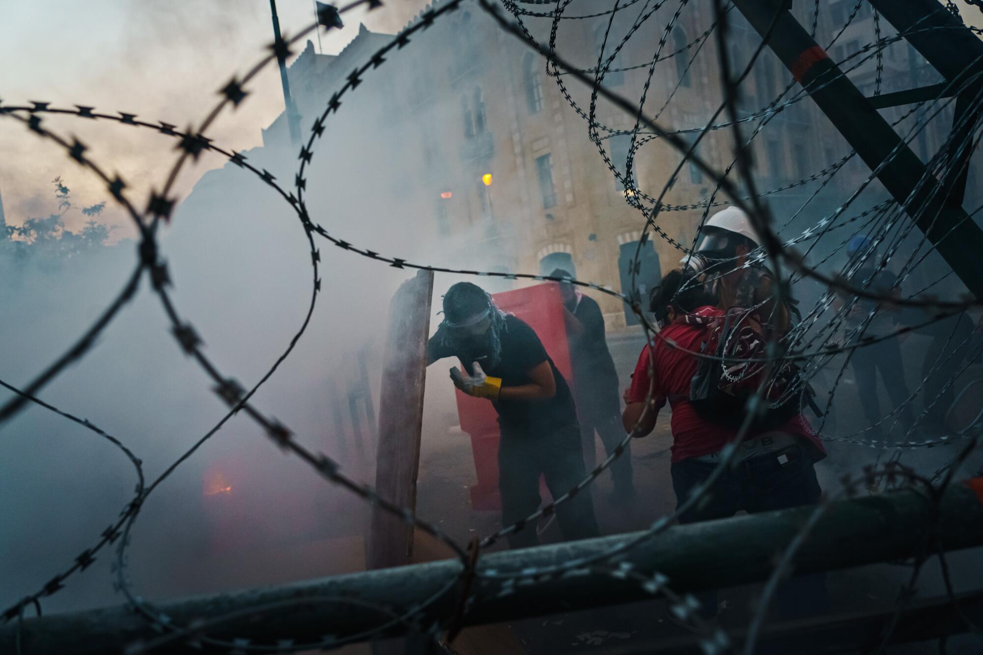 Seen through barbed wire, people, some wearing gas masks, stand amid a haze on a city street.