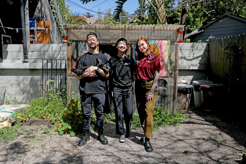 Three people, the one on the left holding a black cat, stand together smiling on a patio