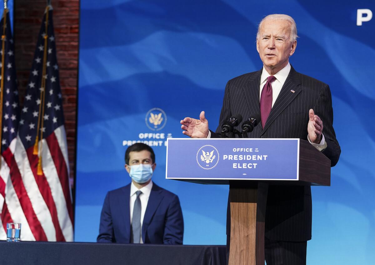 Pete Buttigieg, left, listens as President-elect Joe Biden speaks at a lectern