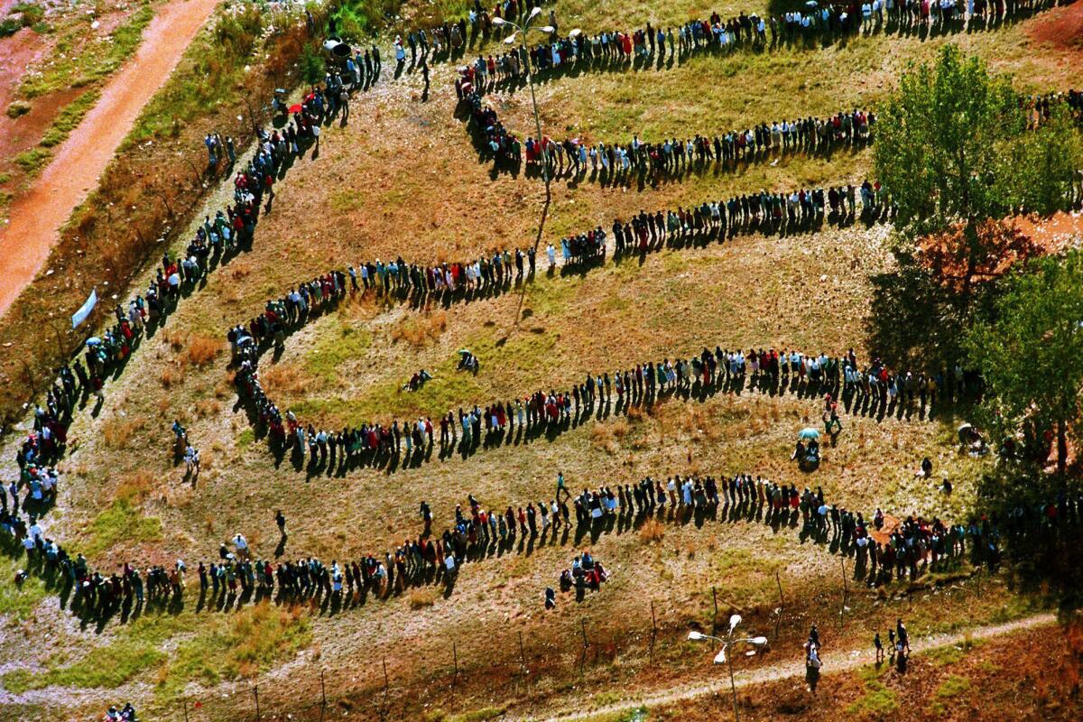 People queue in a long snaking line acrosd a field in an image from above. 