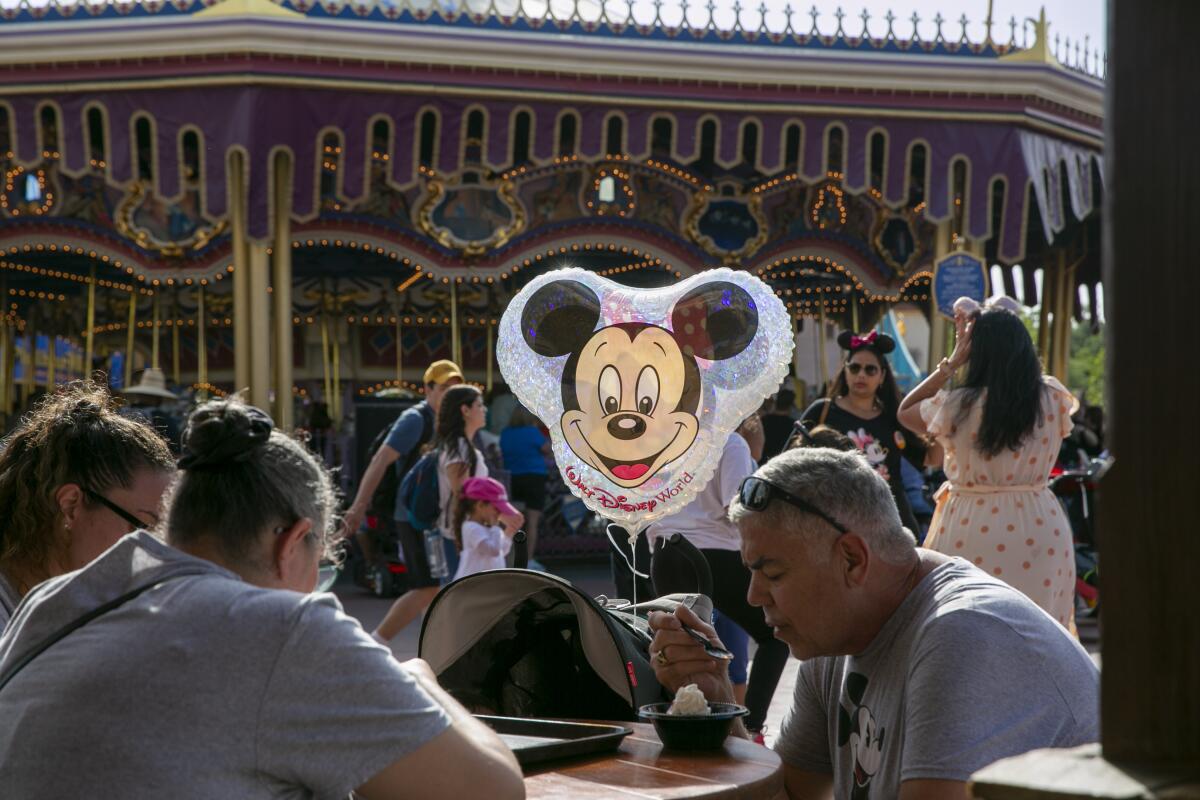 People sit and stand near a merry-go-round and a balloon with Mickey Mouse's face floats among them.