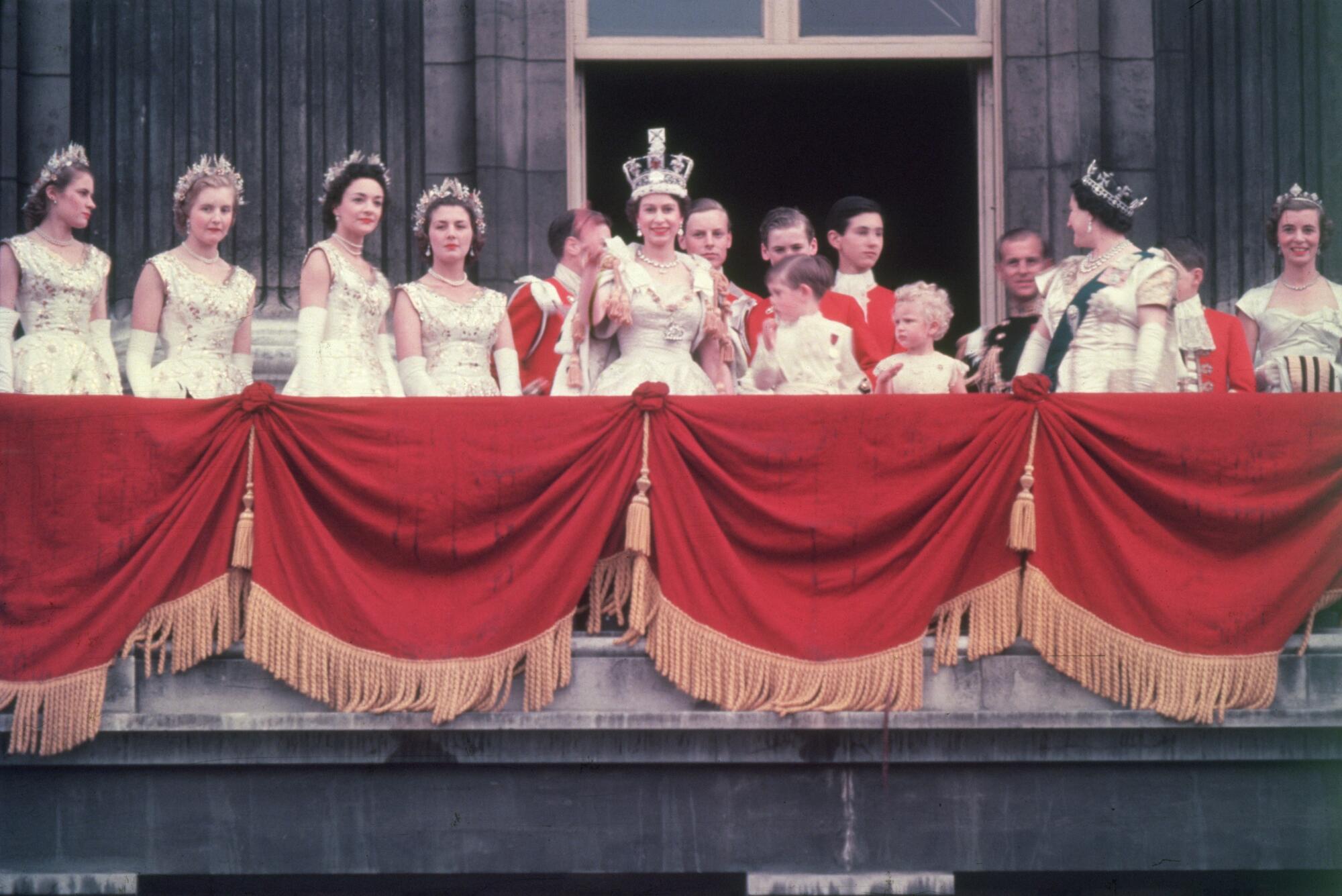 The newly crowned Queen Elizabeth II waves to the crowd from a balcony while several people stand behind her in formal wear. 