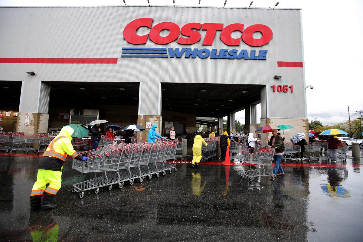 Workers in rain jackets push stacks of shopping carts as customers with umbrellas stand outside a Costco store
