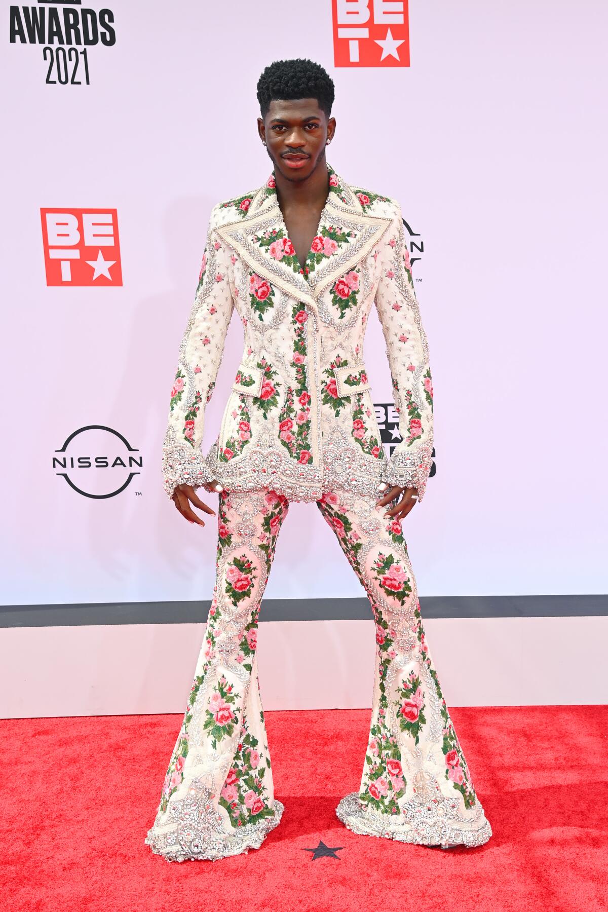 A man in formal attire with pink and red flowers and lace on the white bell-bottomed suit on a red carpet