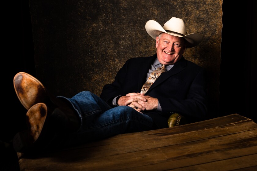 A man in a cowboy hat, jeans and a jacket with a tie smiles as he leans back in a chair with his feet resting on a table.