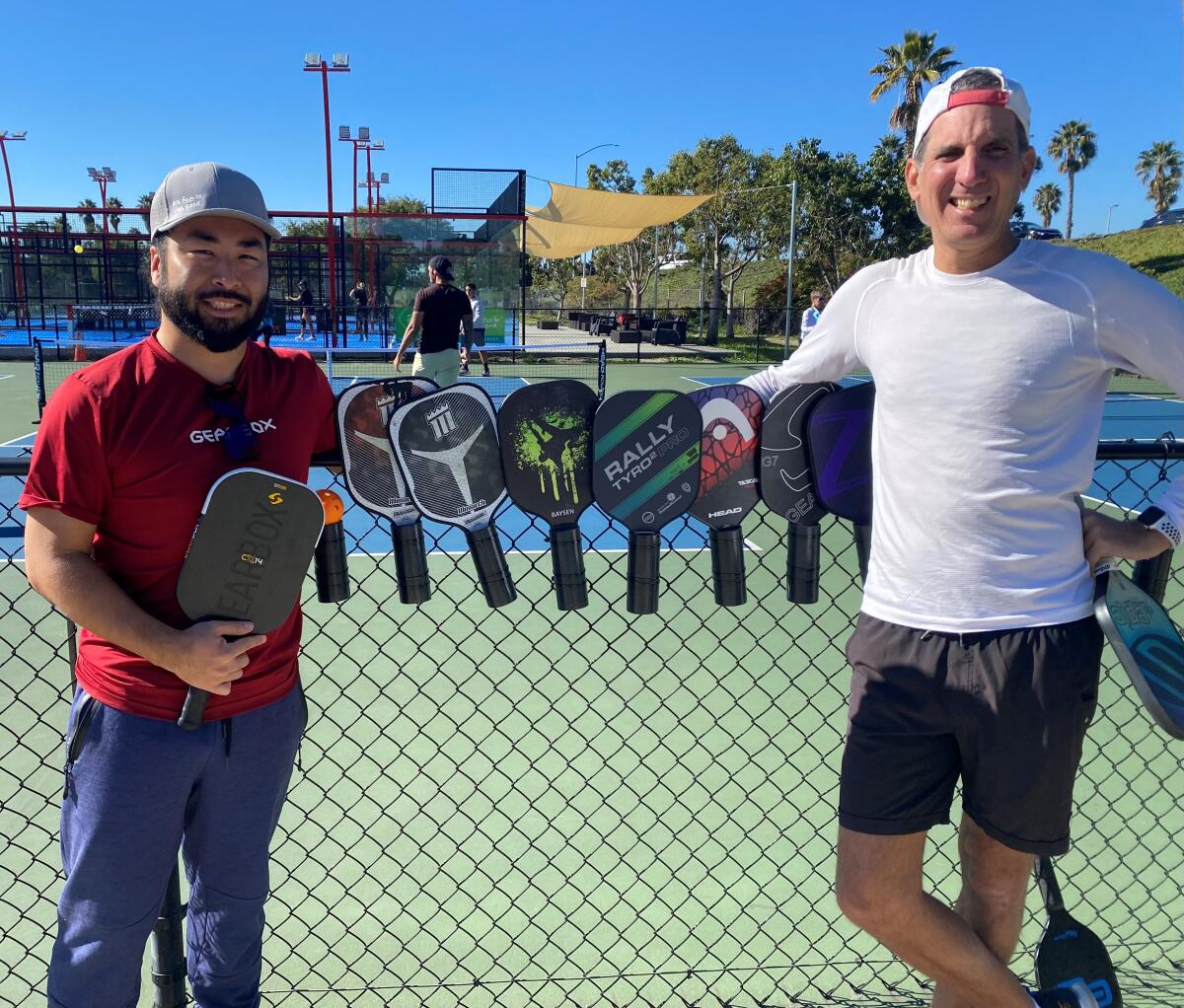 Pickleball instructors and advocates Mike Shinzaki and Stefan Boyland  lean on a fence displaying pickleball paddles