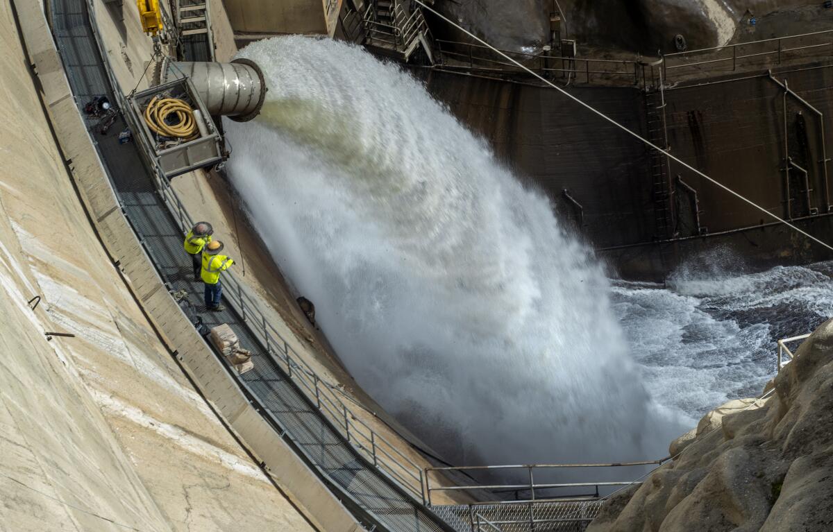 A pair of dam workers in bright yellow jackets watch water cascading from a release valve into a river.
