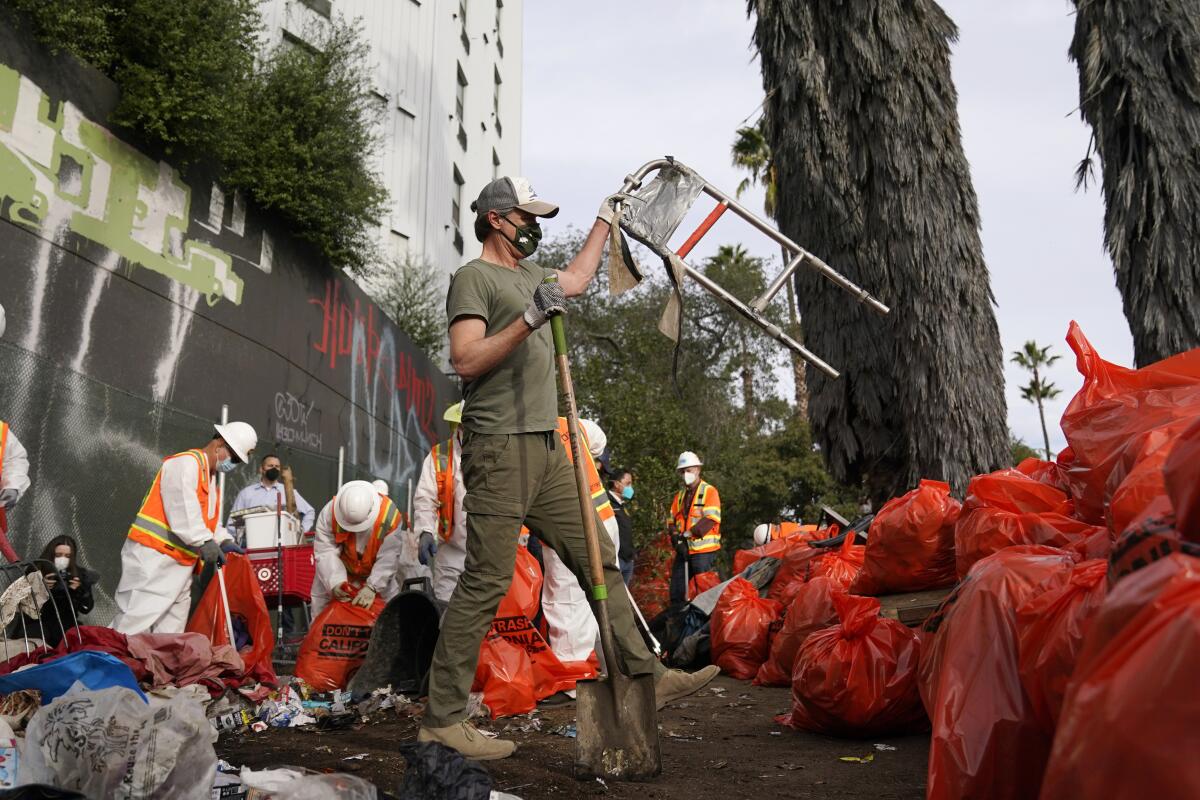 El gobernador de California, Gavin Newsom (c) ayuda a limpiar un campamento de indigentes