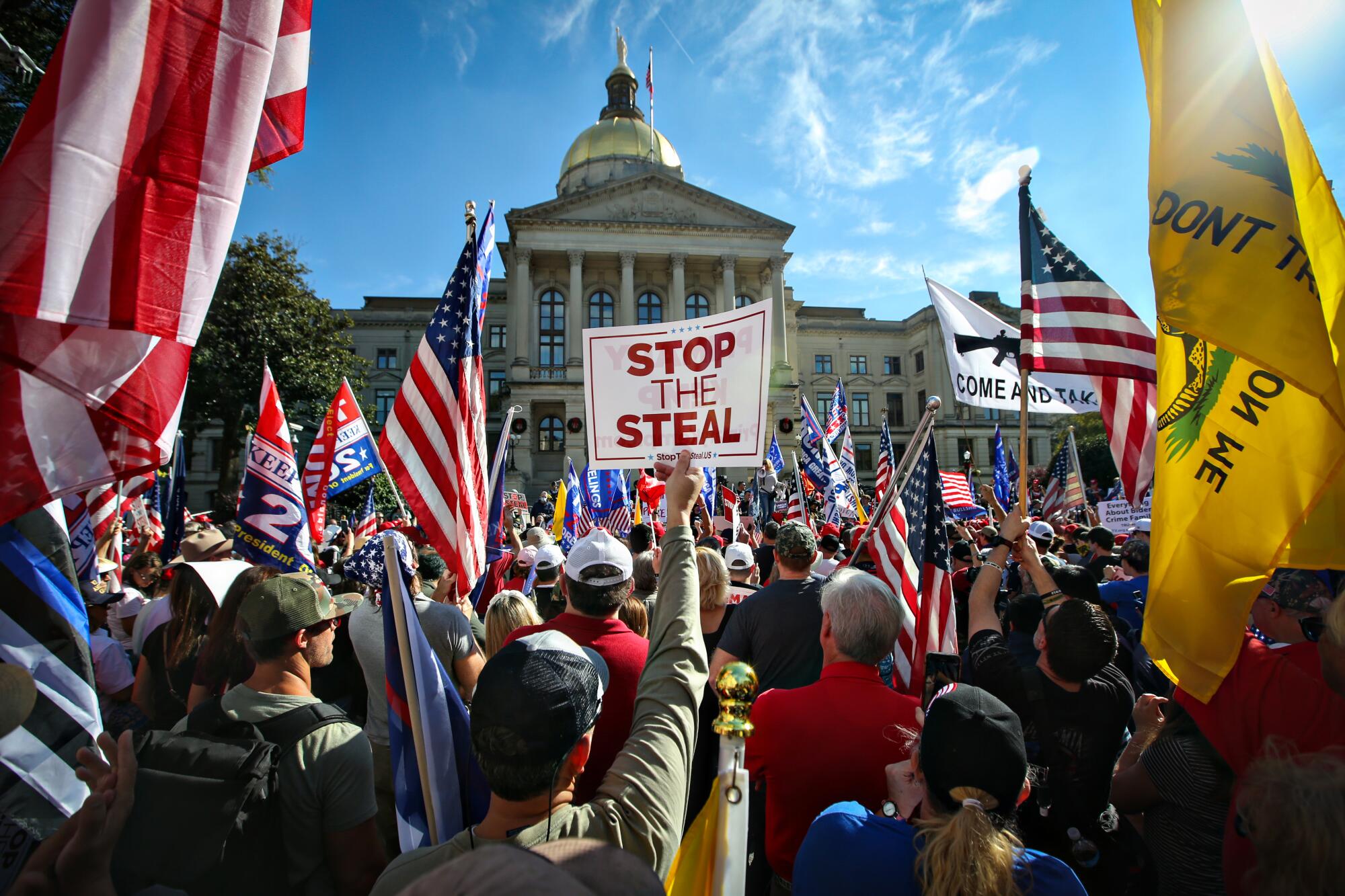 A crowd holding U.S. flags and a sign that says "Stop the Steal Trump" gather in front of a building with pillars and a dome
