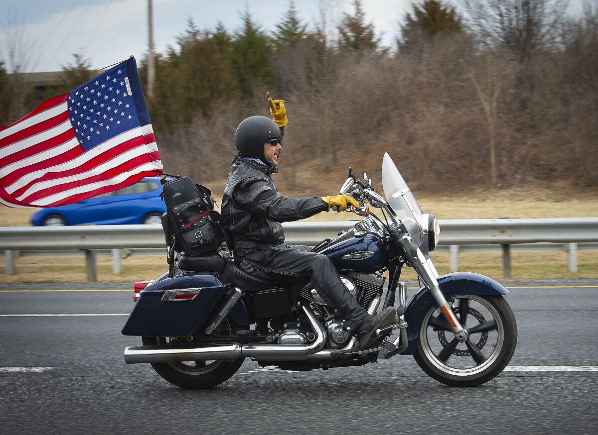 A person on a motorcycle holds up two fingers and a U.S. flag flies behind him on the road. 