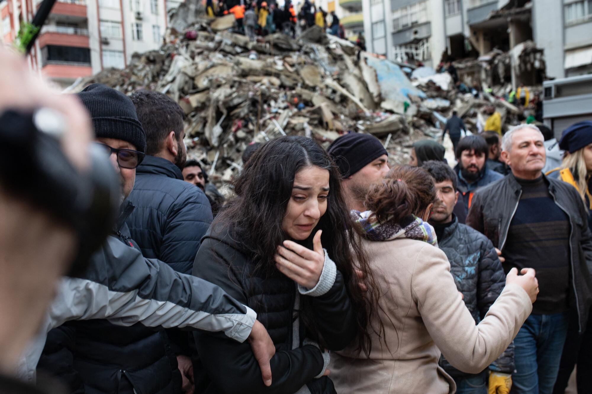 A woman raises her hand to her face, crying, as rescuers search for survivors through the rubble of a building.