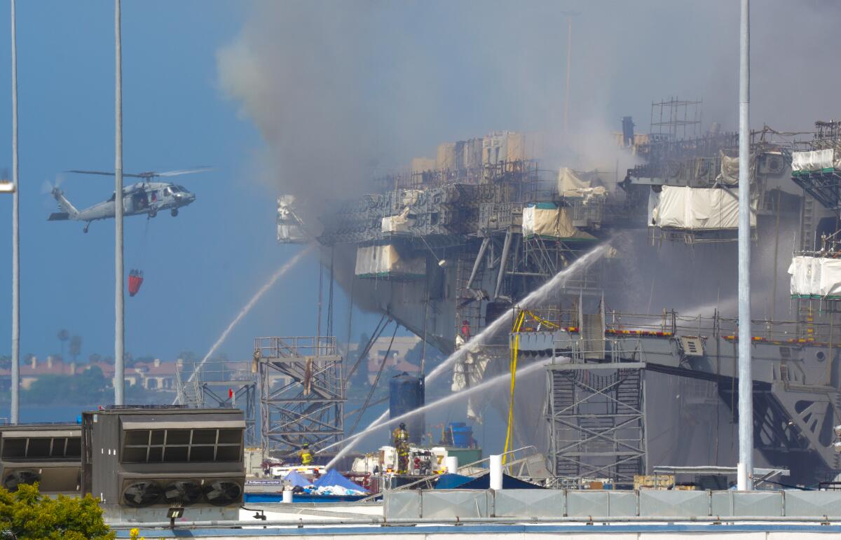 Crews spray water onto a burning Navy warship with smoke coming out of it as a helicopter flies past.