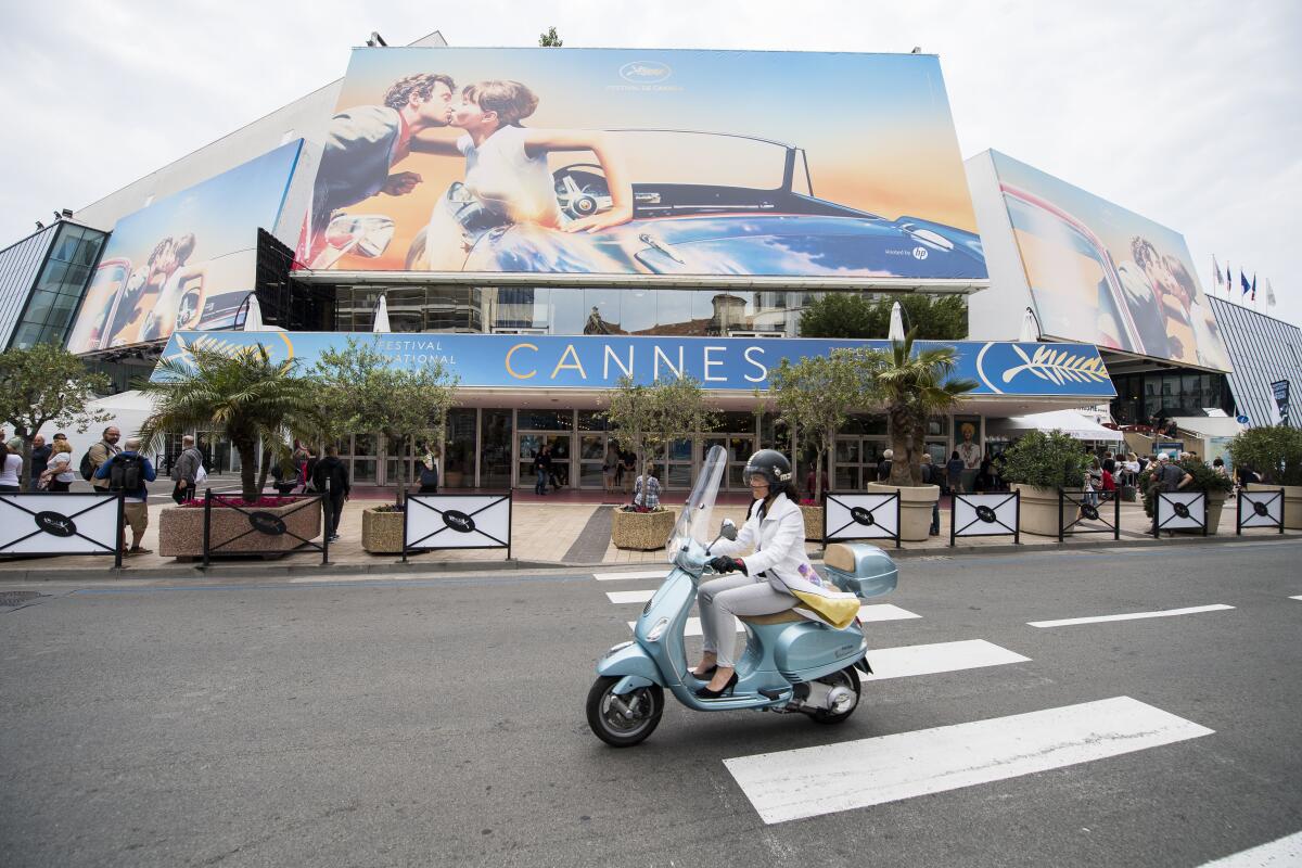 A woman in heels and gloves drives a moped past a theater with a "Cannes" sign.
