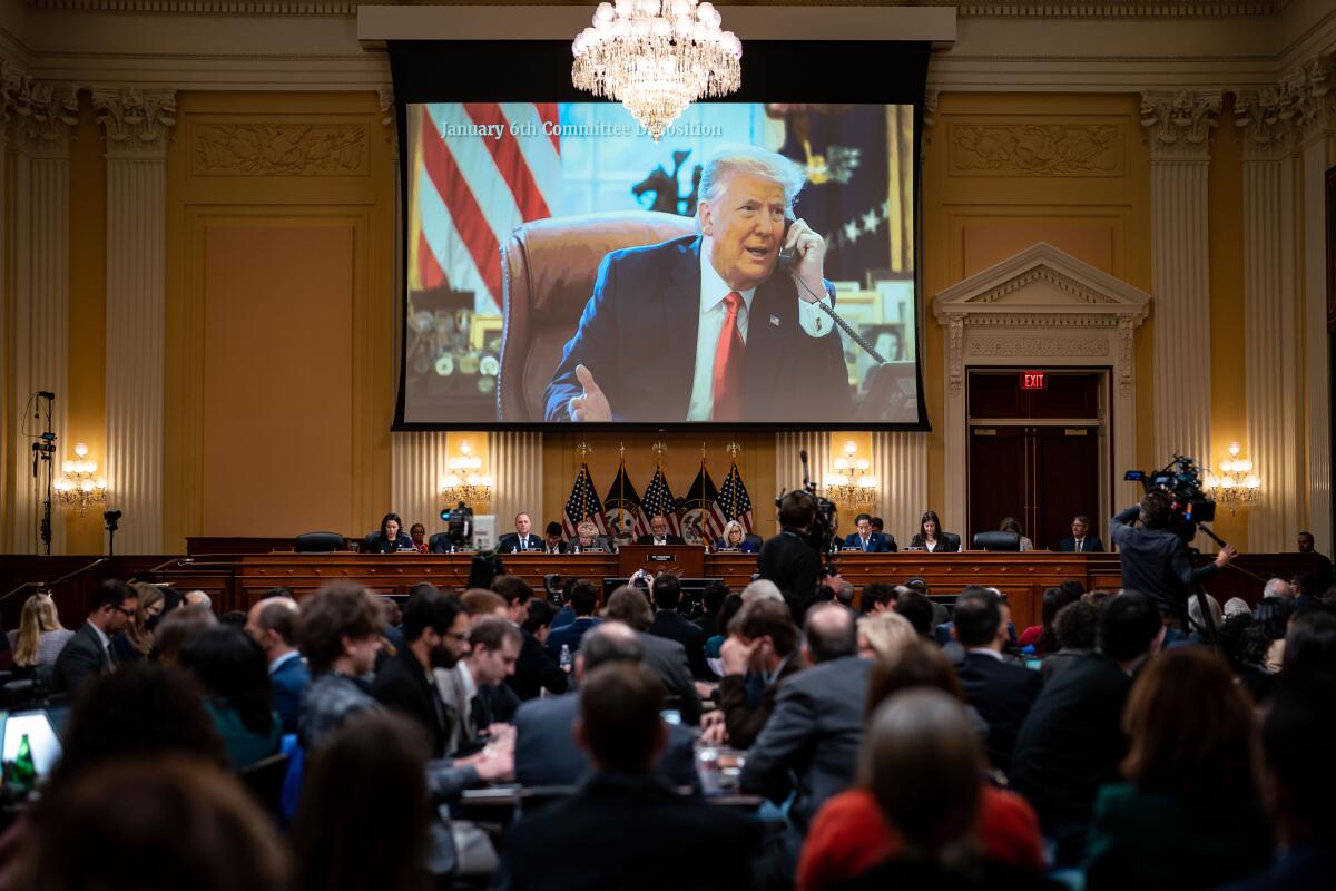 An image of former President Trump talking on the phone is shown on a large screen in a crowded hearing room