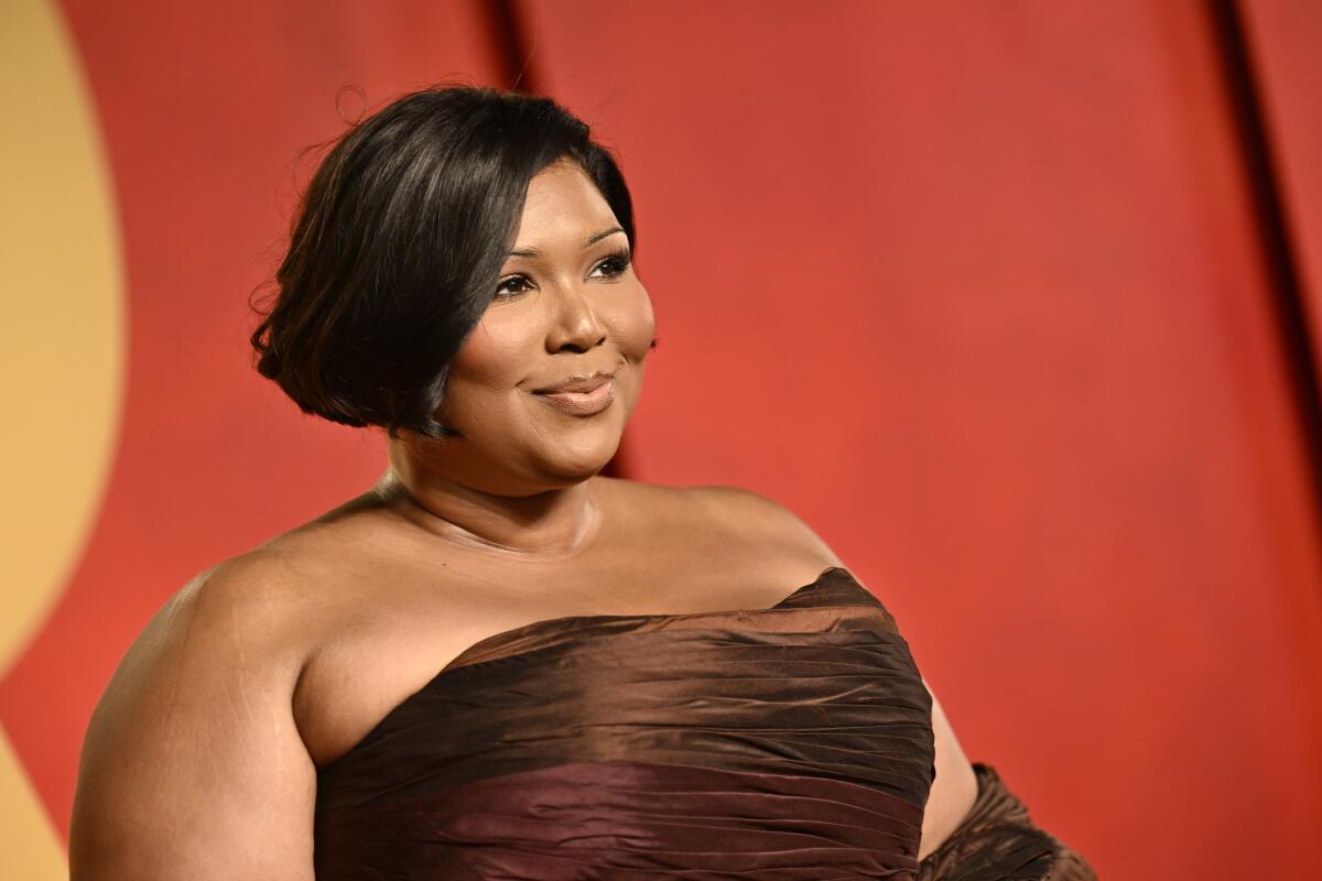Lizzo with her hair in an updo, wearing a brown strapless gown and gloves and posing against a burnt red backdrop