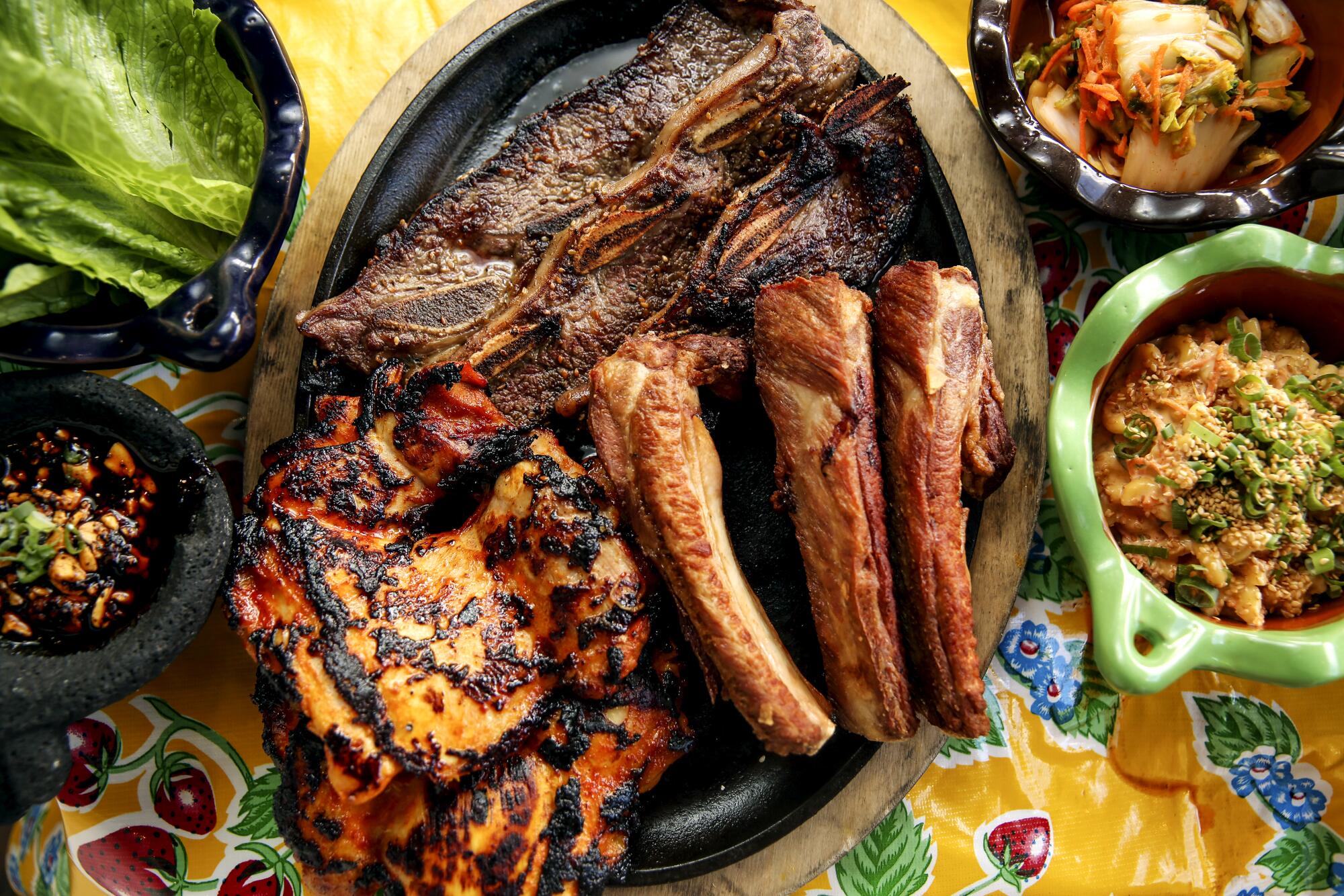 An overhead view of marinated short ribs, spicy chicken thighs, charcoal pork ribs and an assortment of garnishes on a table