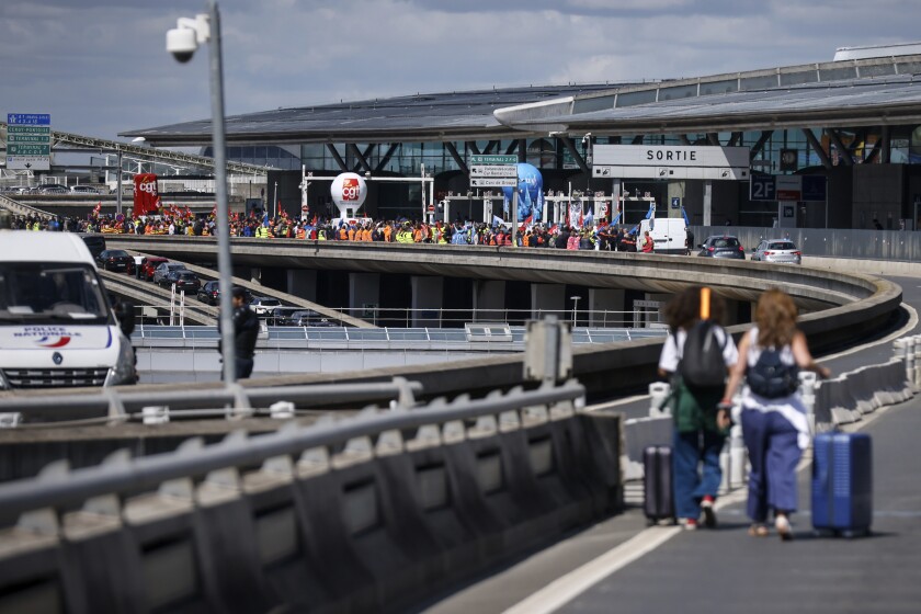 Travelers arrive on foot at Roissy-Charles de Gaulle airport while airport workers demonstrate, left, Friday, July 1, 2022 at Roissy airport, north of Paris. Flights from Roissy-Charles de Gaulle airport in Paris and other French airports faced disruptions Friday as airport workers held a strike and protests to demand salary hikes to keep up with inflation. It's the latest trouble to hit global airports this summer, as travel resurges after two years of virus restrictions. (AP Photo/ Thomas Padilla)