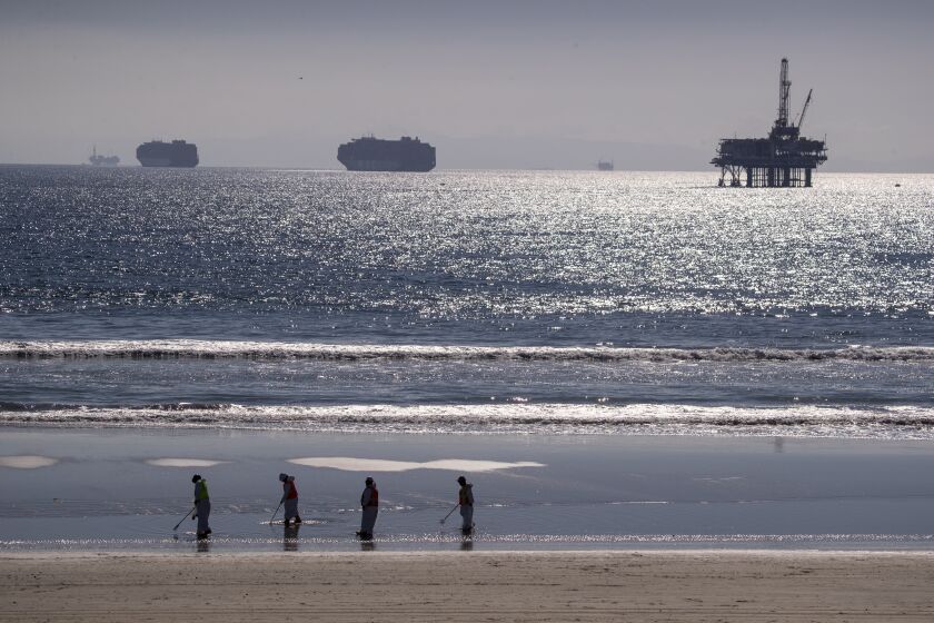 Huntington Beach, CA - October 05: Container ships and an oil derrick line the horizon as environmental oil spill cleanup crews search the beach, cleaning up oil chucks from a major oil spill in Huntington Beach Tuesday, Oct. 5, 2021. Environmental cleanup crews are spreading out across Huntington Beach and Newport Beach to cleanup the damage from a major oil spill off the Orange County coast that left crude spoiling beaches, killing fish and birds and threatening local wetlands. The oil slick is believed to have originated from a pipeline leak, pouring 126,000 gallons into the coastal waters and seeping into the Talbert Marsh as lifeguards deployed floating barriers known as booms to try to stop further incursion, said Jennifer Carey, Huntington Beach city spokesperson. At sunrise Sunday, oil was on the sand in some parts of Huntington Beach with slicks visible in the ocean as well. "We classify this as a major spill, and it is a high priority to us to mitigate any environmental concerns," Carey said. "It's all hands on deck." (Allen J. Schaben / Los Angeles Times)