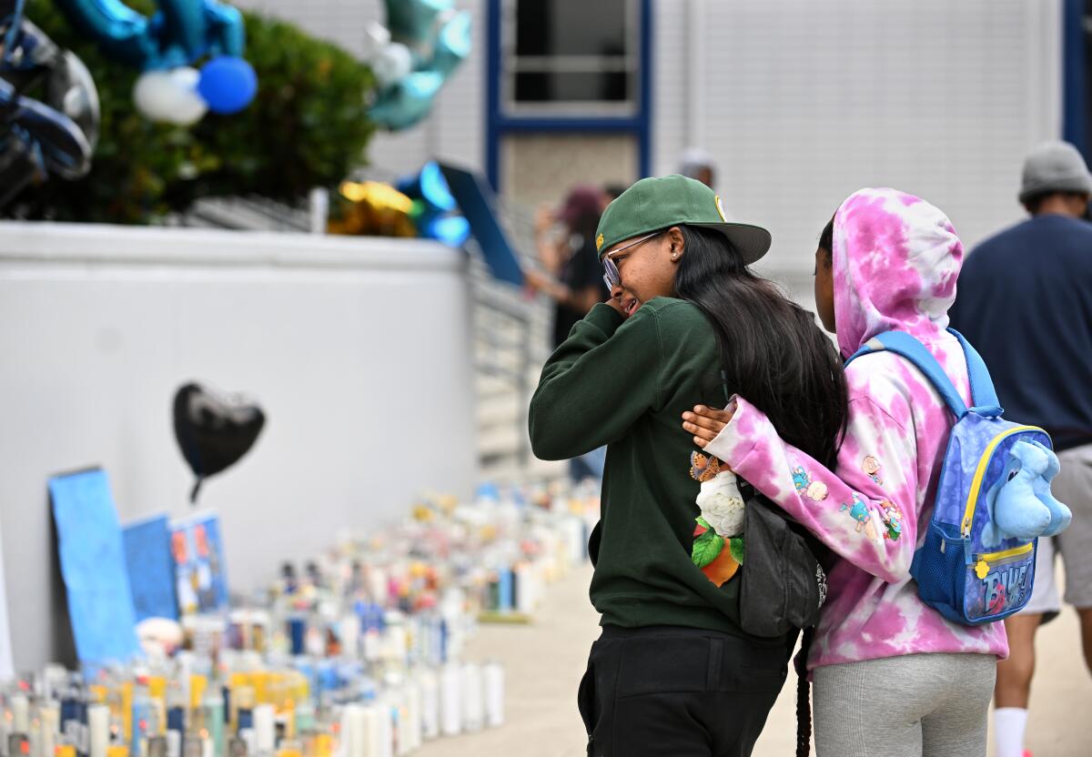 A girl puts her arm around a girl wiping tears from her eyes in front of a memorial with candles, balloons and signs.