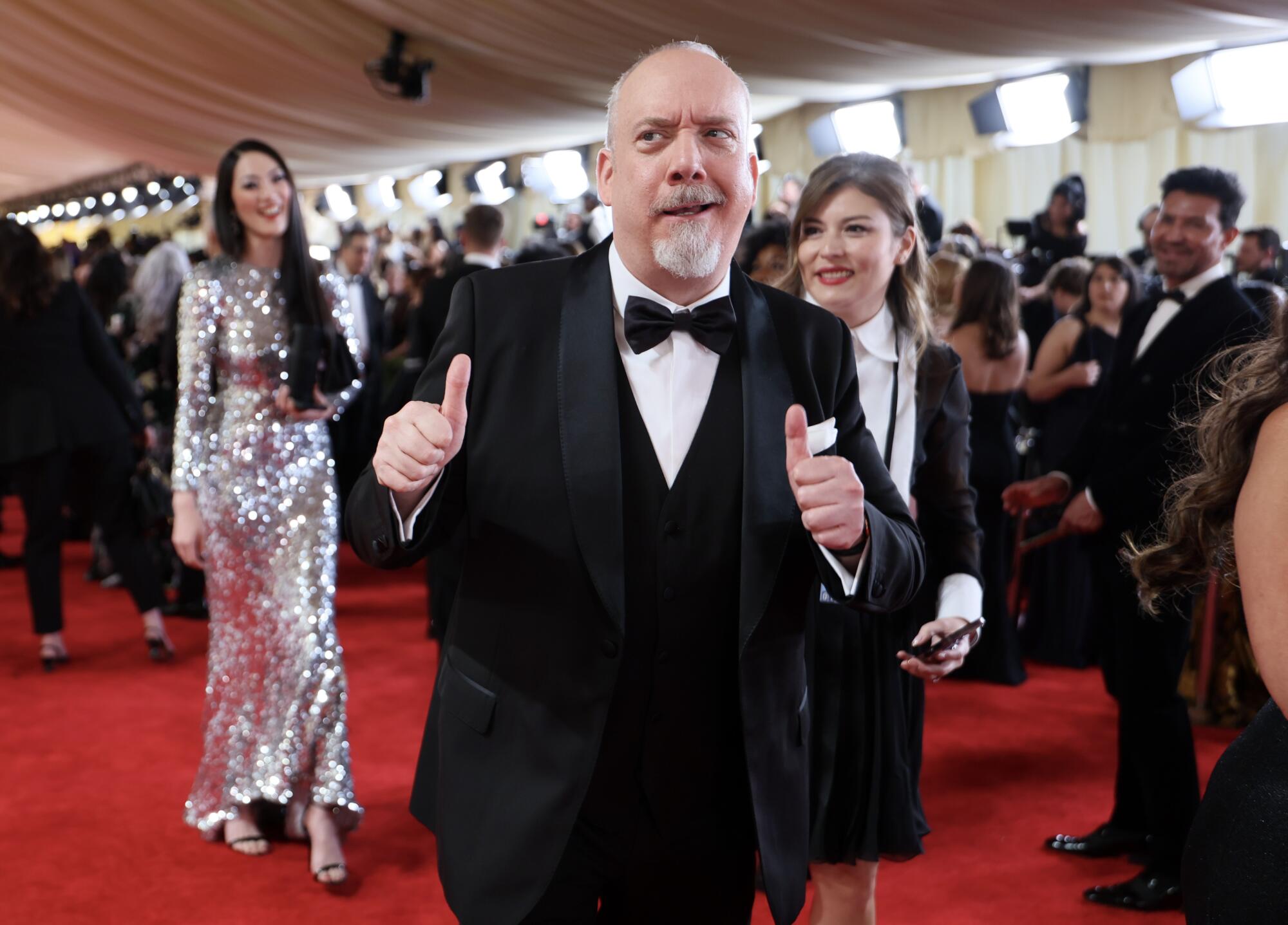 A tuxedoed Paul Giamatti holds up two thumbs as he walks the red carpet. 