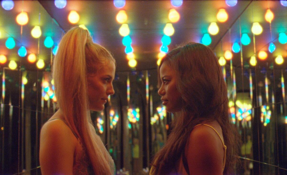 Two young women with long hair are seen in profile staring at each other with lights in the background