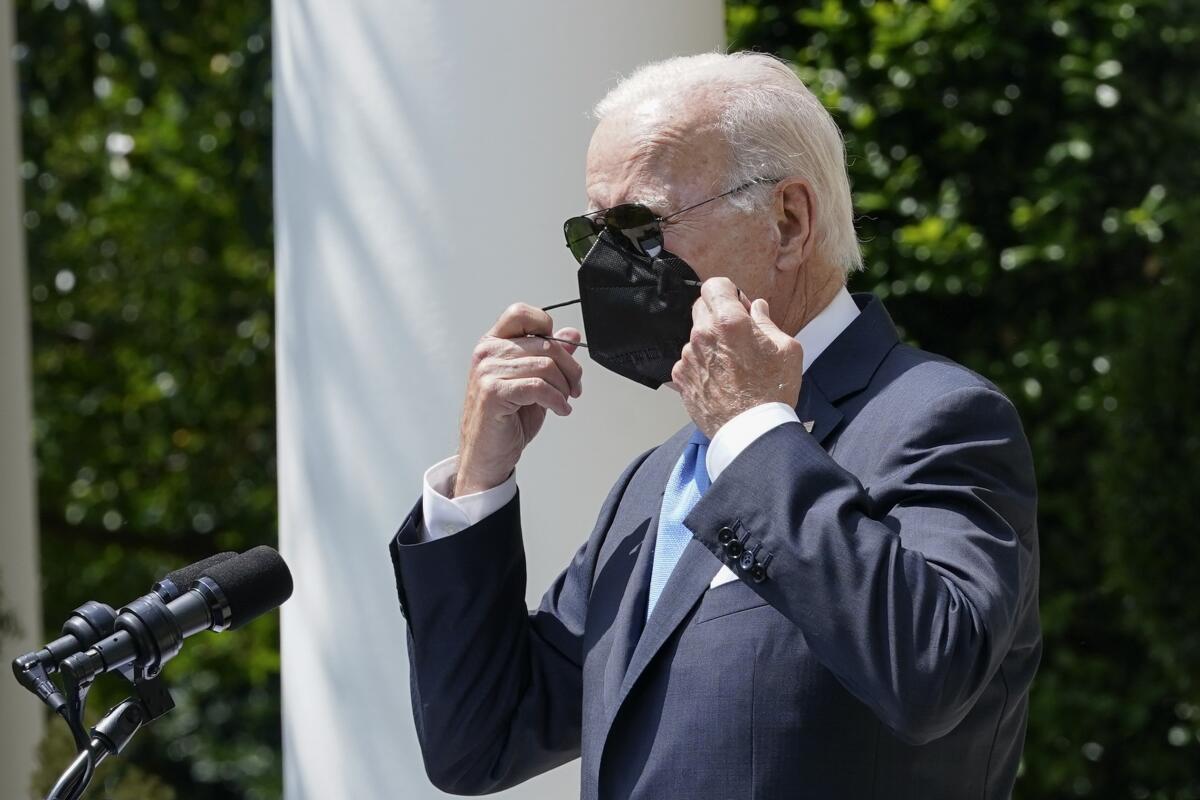 A man with gray hair, wearing sunglasses and a dark suit and light blue tie, removes a black mask before microphones