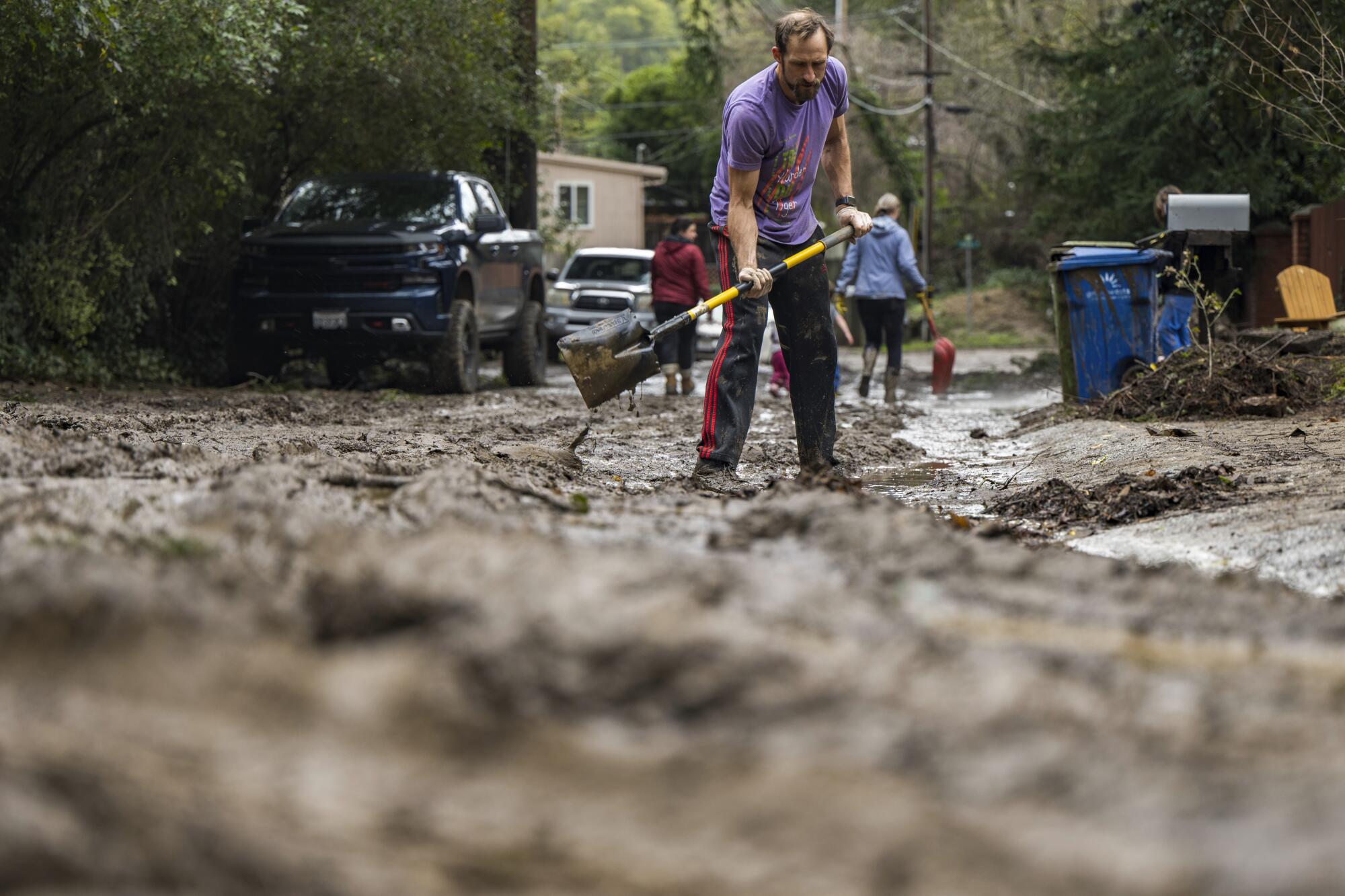 In the center of the frame man shovels mud while two other people walk with shovels in the distance. 