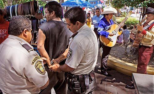 Amid vendors and musicians in the tree-lined central plaza, police in Tecate, Mexico, handcuff a man on unspecified charges. Last December, deputy commander Juan Jose Soriano was assassinated after he reported a cross-border smuggling tunnel. Some suspect the police force has been corrupted by drug lords. More photos >>>
