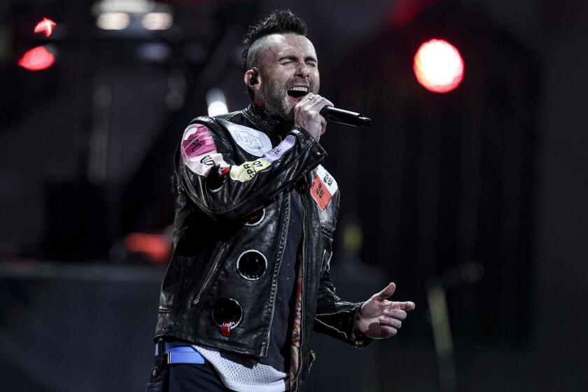Adam Levine wearing a leather jacket with patches and singing into a microphone on stage