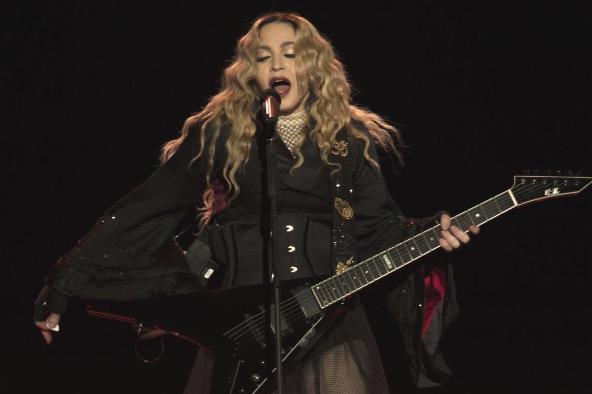 Madonna wears black while playing guitar and singing onstage into a standing microphone