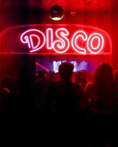 A dark dance club is illuminated by a red and white neon sign that says "disco"