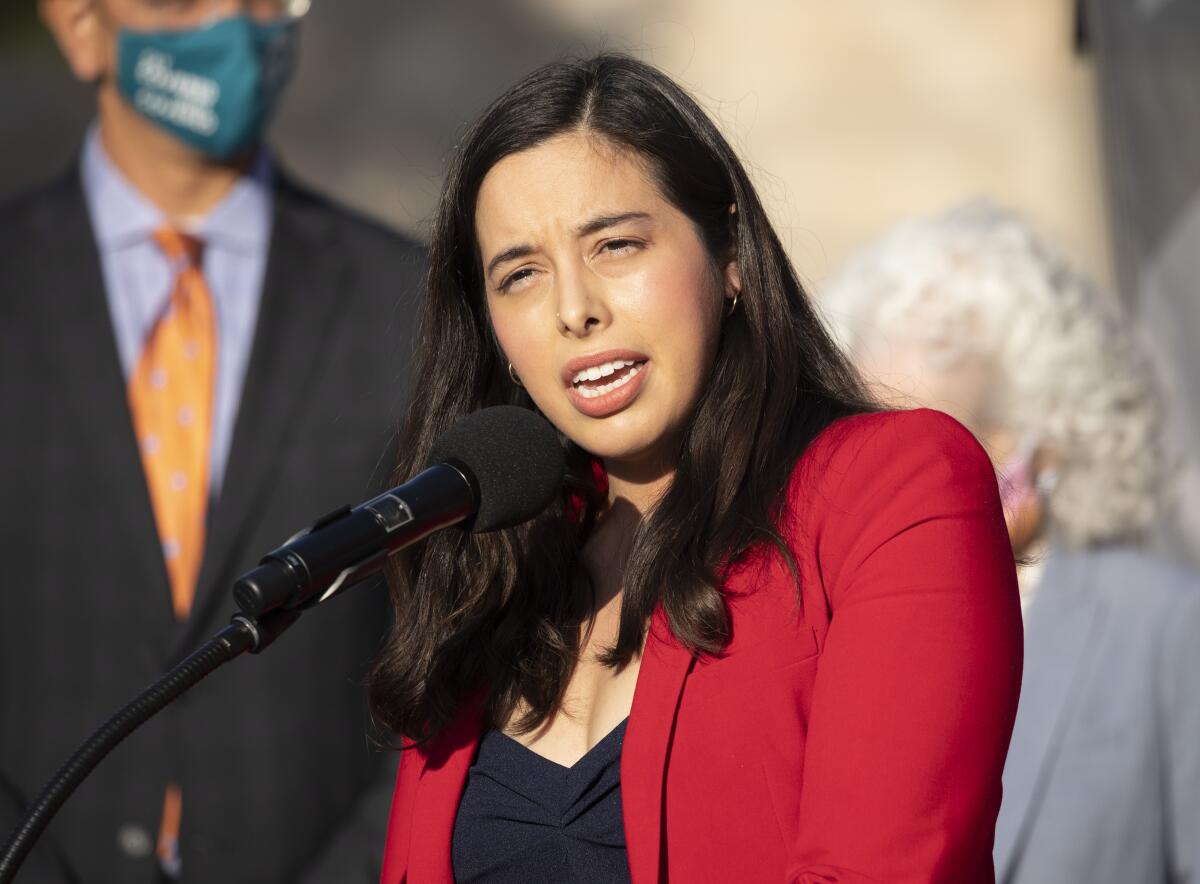 A woman in a red jacket speaks into a microphone at a lectern.