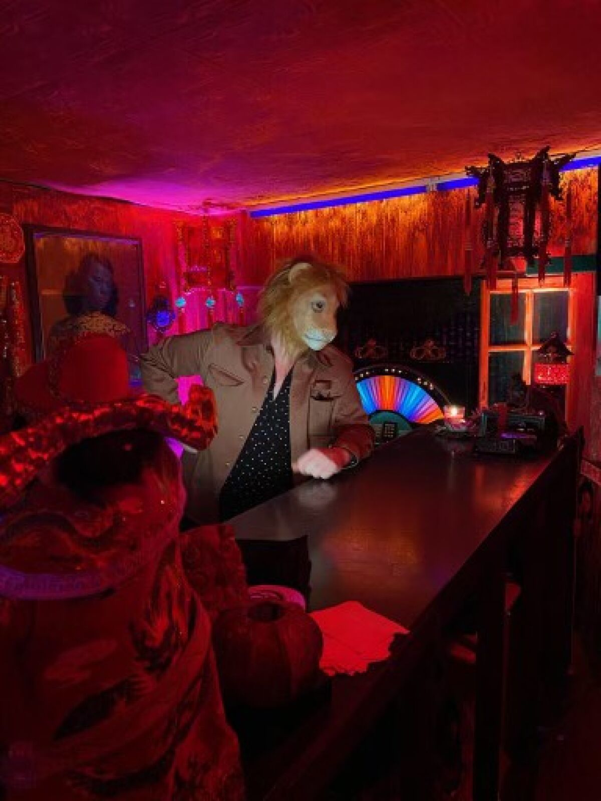 A guy wearing a lion-head mask stands at the bar in a red-lighted club.