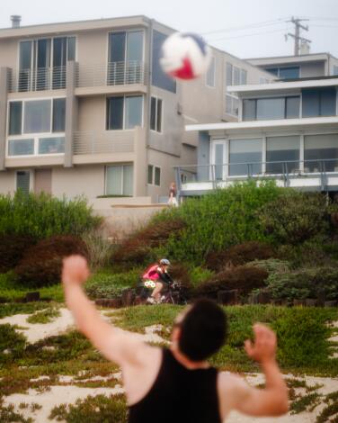 A person about to hit a tossed-up ball in front of beachfront houses.