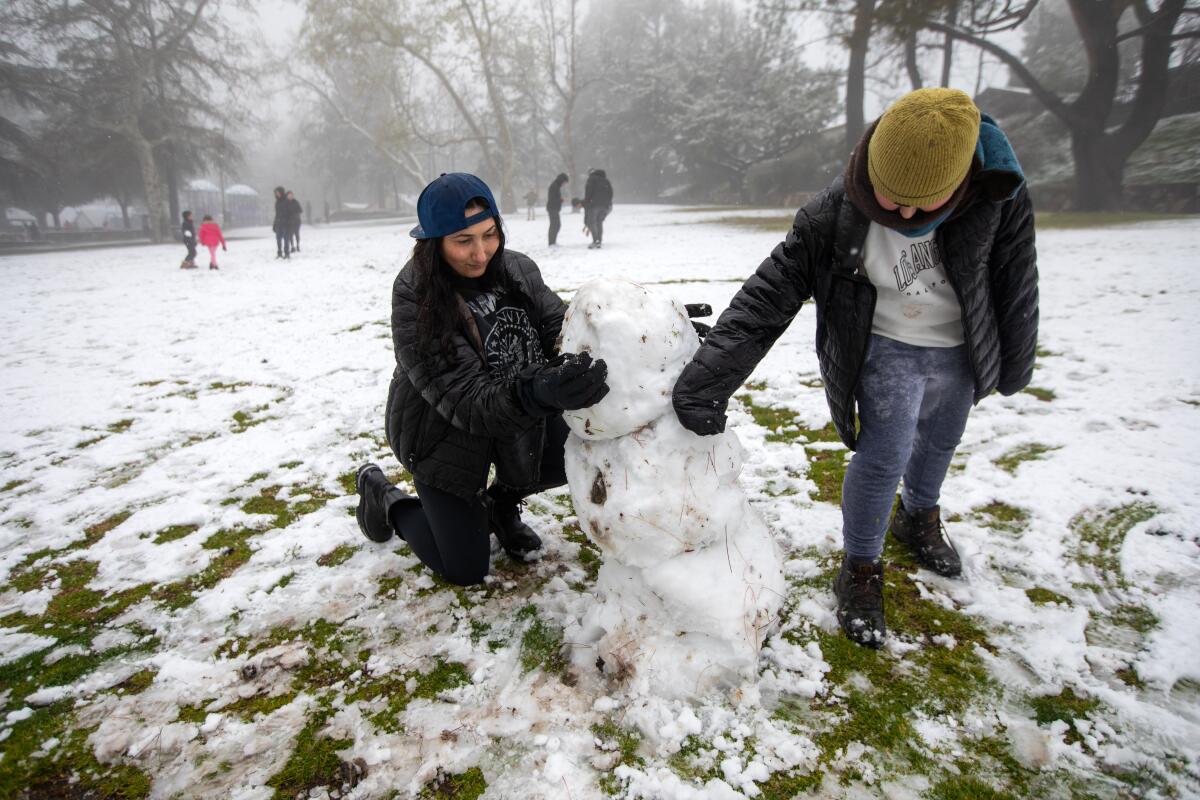 A woman crouches and supports a snowman in a snowy field as a child adds more snow