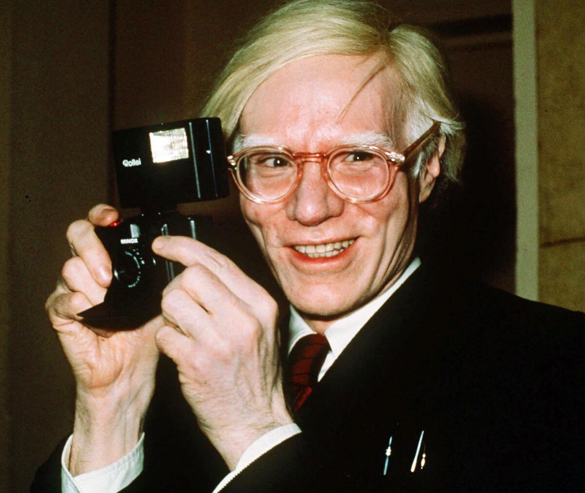 A vintage photo from 1976 shows Andy Warhol smiling brightly and holding a camera. 