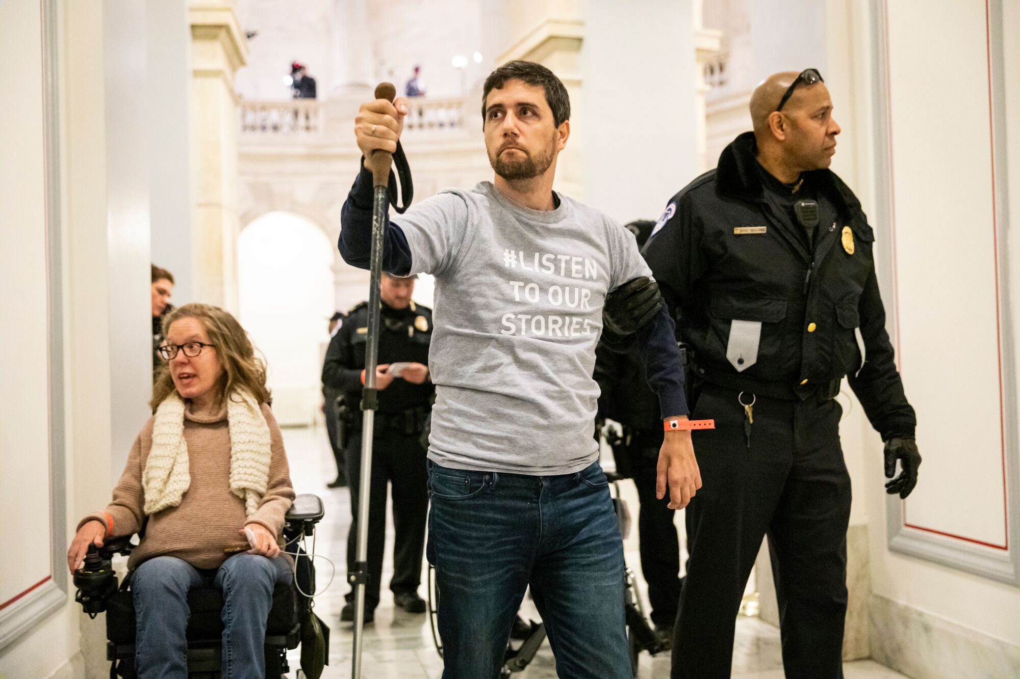 A woman in a wheelchair, a man walking with a hiking pole and several security officers.