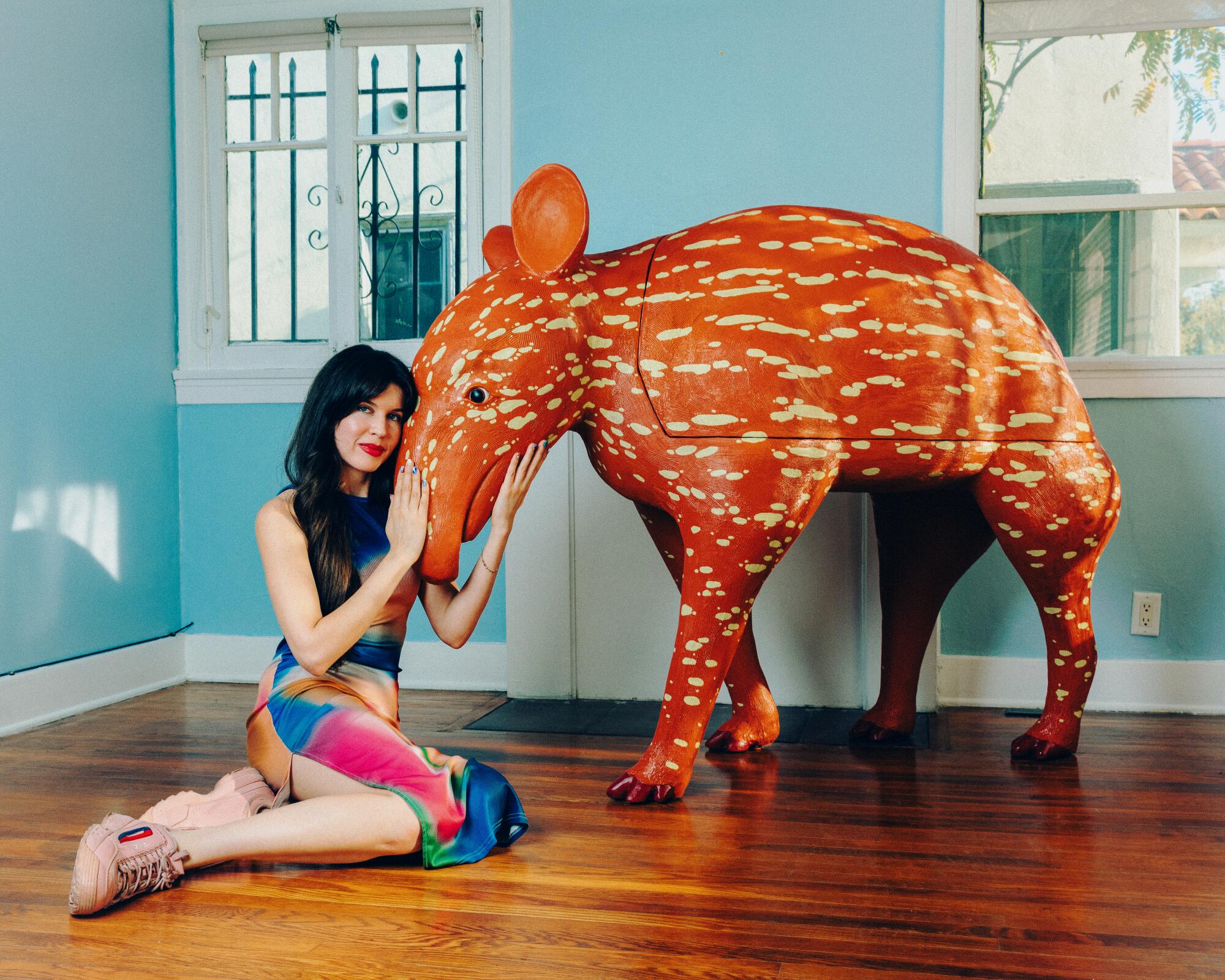 A portrait of a woman with dark hair and colorful dress, sitting on the floor nose to nose with a tapir sculpture.