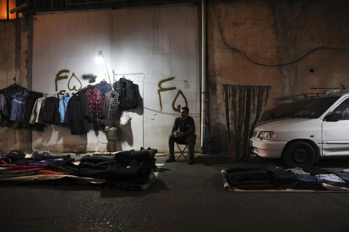 A vendor sits by a rack of clothes on a dark street in Tehran.