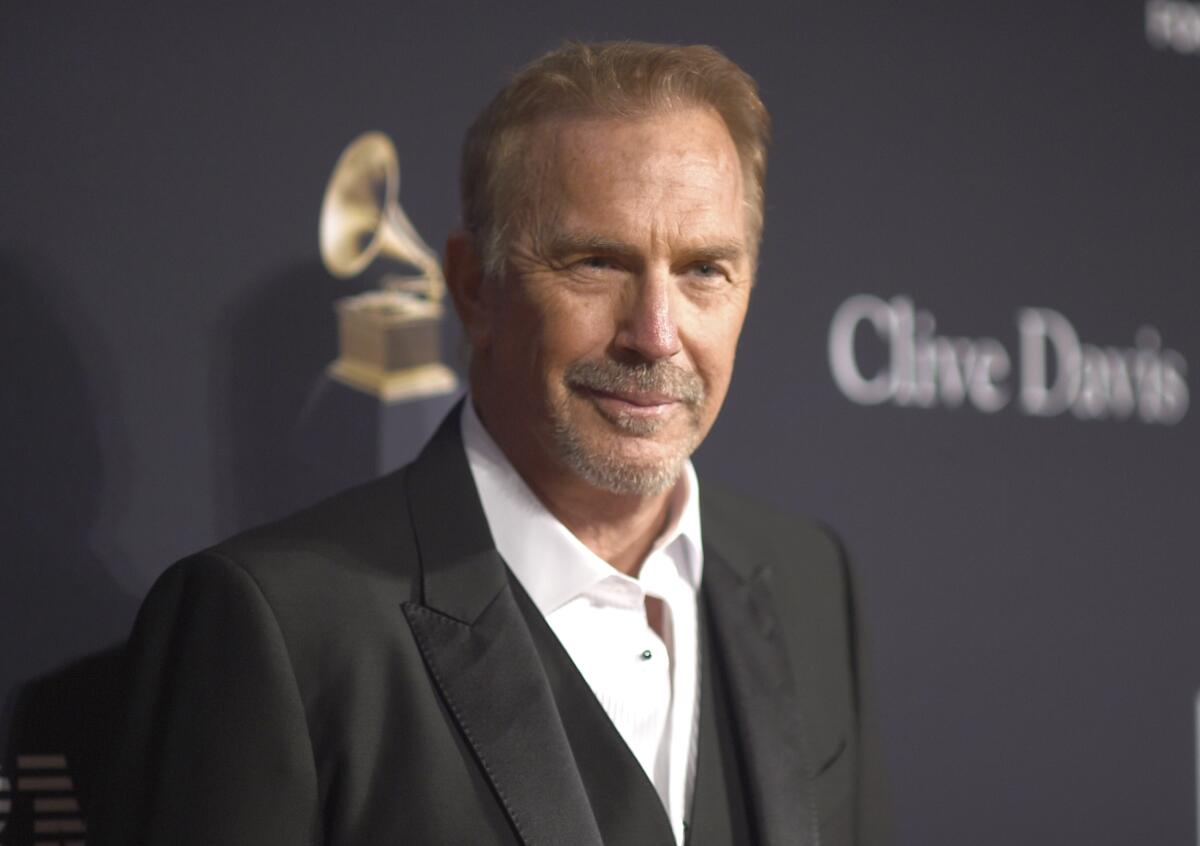 Kevin Costner posing at an angle in a black blazer and vest and white suit shirt